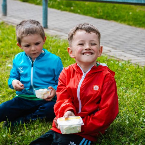 7. Zwei glückliche Kindergartenkinder sitzen am Spielfeldrand im Gras und genießen ihr Eis von Station 57 zur Abkühlung.
