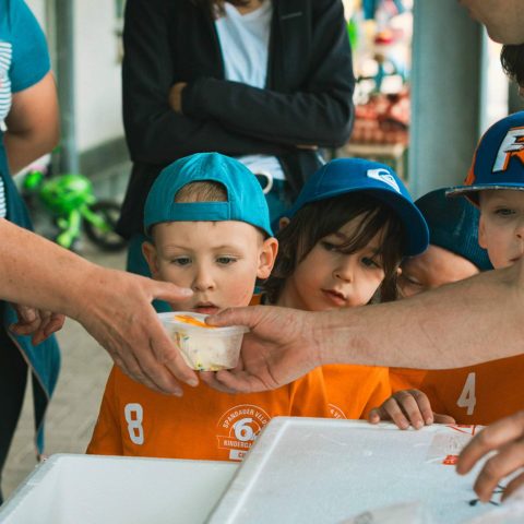 8. Kinder in orangefarbenen Trikots warten gespannt am Verpflegungsstand und bekommen Becher mit Eis gereicht.