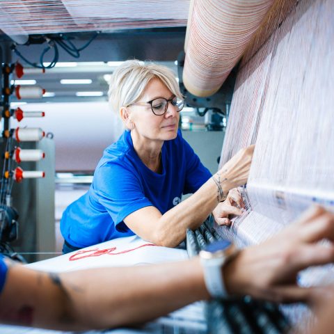 Eine blonde Mitarbeiterin im blauen T-Shirt beugt sich über eine große industrielle Webmaschine. Sie prüft mit den Händen präzise die unzähligen, fein aufgespannten Kettfäden beim Vorrichten für die Veloursherstellung. Im Vordergrund sind unscharf die Arme einer Kollegin zu sehen.