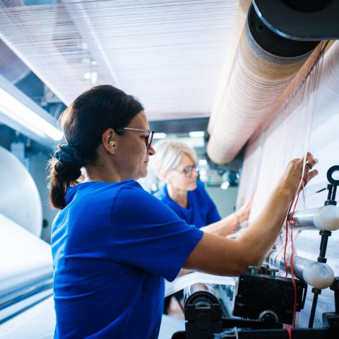 Seitenansicht in der Produktionshalle: Eine dunkelhaarige Mitarbeiterin mit Brille und gelbem Gehörschutz arbeitet konzentriert an einer großen Kettbaum- und Vorrichtmaschine. Sie greift präzise nach den feinen Fäden. Im Hintergrund ist eine weitere Kollegin an der Maschine erkennbar.