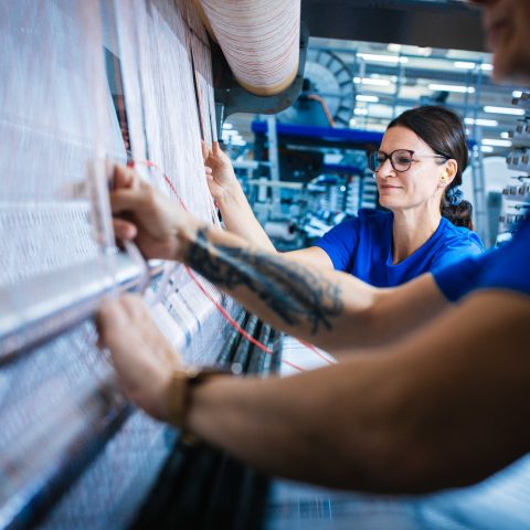 Perspektive entlang der Maschine: Eine Mitarbeiterin mit Brille blickt konzentriert auf die aufgespannten Fäden, während im Vordergrund der tätowierte Arm einer Kollegin zu sehen ist, die die Fäden millimetergenau ausrichtet.