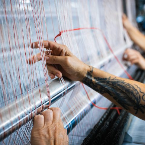 Eine Nahaufnahme, die das Herzstück der Vorbereitung zeigt: Ein tätowierter Unterarm und Hände führen die feinen weißen und roten Kettfäden mit viel Fingerspitzengefühl über die glänzenden Metallstangen einer Webmaschine.