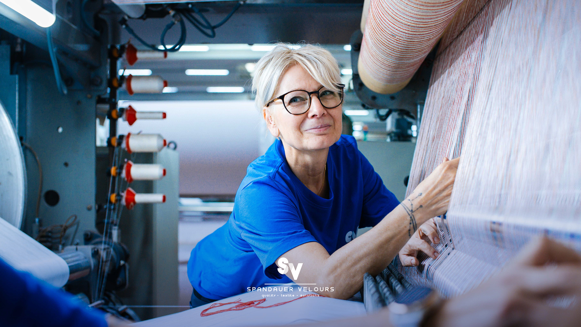 Eine blonde Mitarbeiterin im blauen T-Shirt beugt sich über eine große industrielle Webmaschine. Sie prüft mit den Händen präzise die unzähligen, fein aufgespannten Kettfäden beim Vorrichten für die Veloursherstellung. Im Vordergrund sind unscharf die Arme einer Kollegin zu sehen. Sie lächelt rüber zu ihrer Kollegin. Unten ist mittig das weiße Logo von Spnadauer Velours platziert.