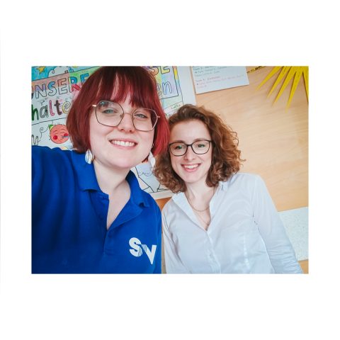 Zwei lächelnde Mitarbeiterinnen der Spandauer Velours im blauen Firmen-Poloshirt und weißer Bluse beim Ausbildungsevent an der Schule.
