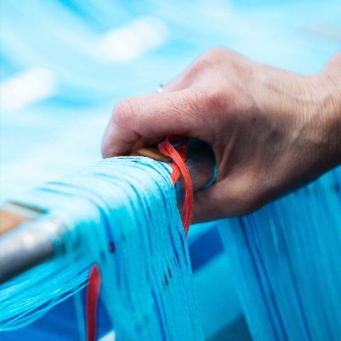 Handarbeit an der Webmaschine: Mitarbeiter führt leuchtend blaue und rote Garnfäden präzise per Hand zusammen.
