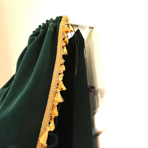 Detailaufnahme des Vorhangstoffs: Der tiefgrüne, weiche Veloursstoff fällt in eleganten Falten. Die Kante ist mit einer filigranen, gelb-goldenen Fransenborte verziiert.