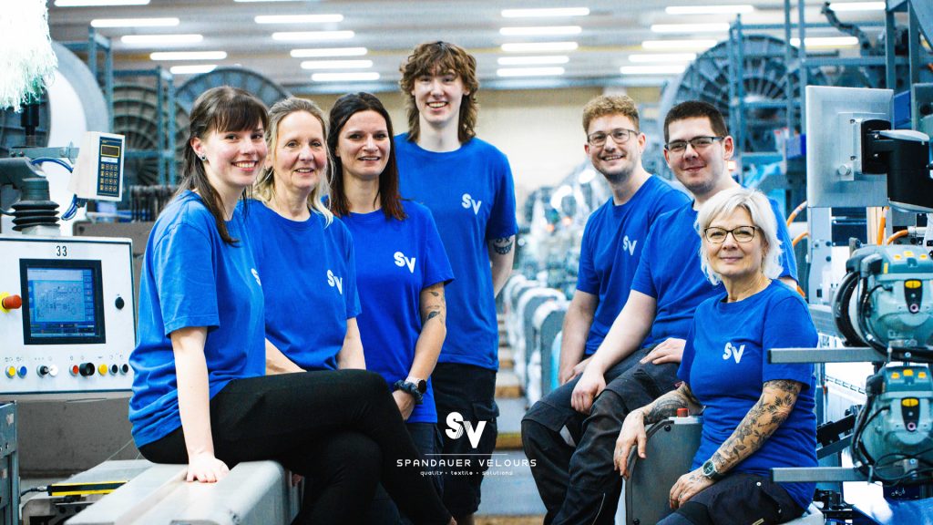 Team von Spandauer Velours sitzt in blauer Arbeitskleidung in der Produktionshalle und lächelt