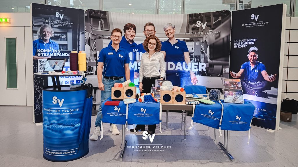 Messestand von Spandauer Velours auf der Ausbildungsmesse in Glauchau mit zwei lächelnden Mitarbeitenden, Messewand mit Aufschrift „Komm ins Team Spandauer“ und präsentierten Textilprodukten sowie Farbwalzen