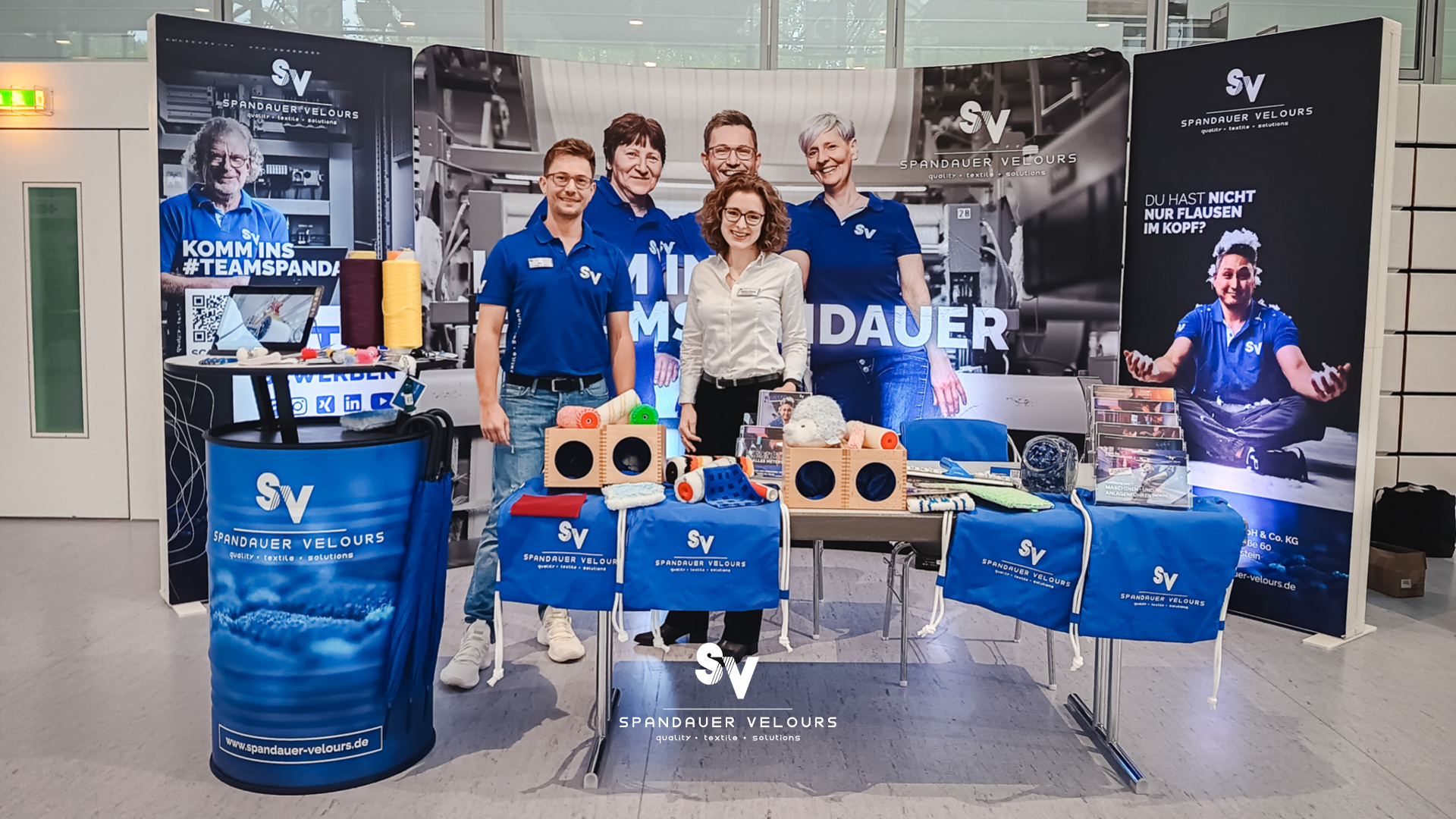 Messestand von Spandauer Velours auf der Ausbildungsmesse in Glauchau mit zwei lächelnden Mitarbeitenden, Messewand mit Aufschrift „Komm ins Team Spandauer“ und präsentierten Textilprodukten sowie Farbwalzen